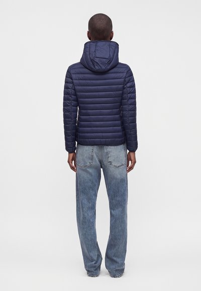 Personne portant une veste matelassée bleu marine avec capuche et un jean bleu clair ample, debout de dos sur un fond uni.
