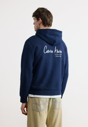Homme portant un sweat à capuche bleu marine avec le logo « Calvin Klein » dans le dos, associé à un pantalon beige, debout devant un fond clair uni.
