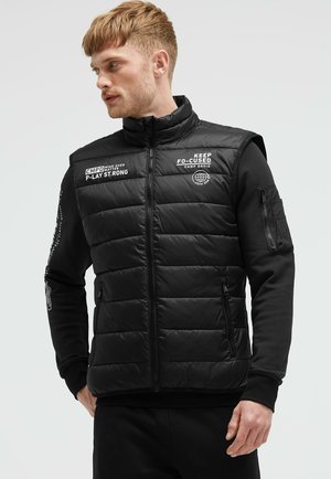 Homme aux cheveux clairs portant une doudoune noire avec un texte blanc sur un t-shirt noir à manches longues, debout devant un fond clair uni, regardant vers la gauche.