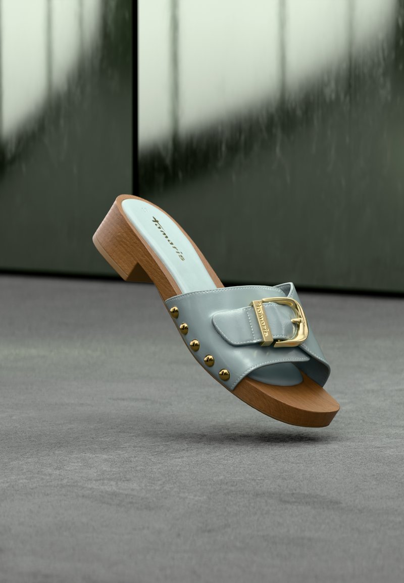 Sandale à glissière en cuir bleu avec semelle en bois, boucle dorée et clous, posée au-dessus d'une surface grise sur un fond sombre flou.