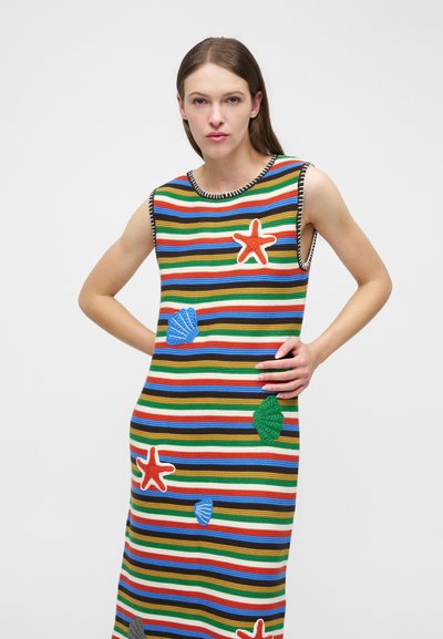 Frau trägt ein ärmelloses, mehrfarbig gestreiftes Kleid mit aufgestickten Seestern- und Muschelapplikationen und posiert mit einer Hand in der Hüfte.