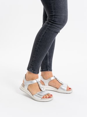 Person wearing silver open-toe sandals with ankle straps and dark skinny jeans, standing on a white surface.