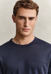 Giovane uomo con capelli castani corti indossa una maglietta blu scuro a girocollo, guarda direttamente la fotocamera con un'espressione neutra.