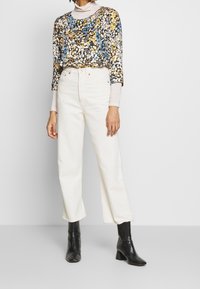 Mønstrede bluse med flerfarvede blomster i rund halsudskæring, parret med højtaljede hvide brede jeans og sorte hælede ankelstøvler.
