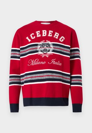 Uomo che indossa occhiali da sole neri, maglione a righe rosse con la scritta "ICEBERG Milano Italia", pantaloni neri e mocassini marrone scuro, in piedi davanti a uno sfondo bianco.