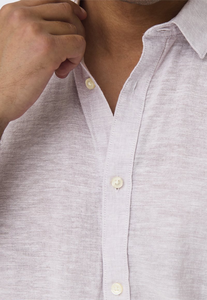 Light pink linen shirt with a button-down collar, featuring small white buttons and a textured fabric. Close-up on the collar and chest area.
