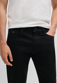 Homme portant un jean noir avec un petit logo BOSS sur la poche, une main dans la poche et l'autre détendue sur le côté, chemise blanche partiellement visible.