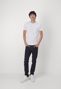 White cotton t-shirt paired with dark blue denim jeans and white sneakers. Model stands with hands in pockets, against a plain background.