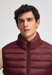 Chaleco acolchado burdeos con cuello alto, cremallera frontal y detalle sutil del logotipo azul en el pecho, fabricado con material ligero y suave.