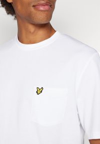 T-shirt bianca in cotone con scollatura rotonda, dotata di tasca sul lato sinistro e dettaglio del logo ricamato giallo.