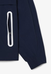 Manche de veste bleu marine avec un poignet élastique et une poche verticale zippée bordée de blanc sur le côté inférieur.