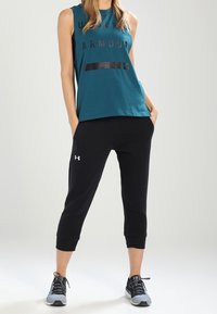Turkos ärmlös topp med "UNDER ARMOUR" i svart, i kombination med svarta capribyxor med uppluckrad nederkant och vitt logotyp på vänster ben.