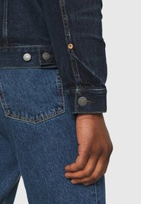 Dark denim jacket with a fitted design, visible stitching, and metal buttons. Worn with blue jeans, showcasing a textured surface.