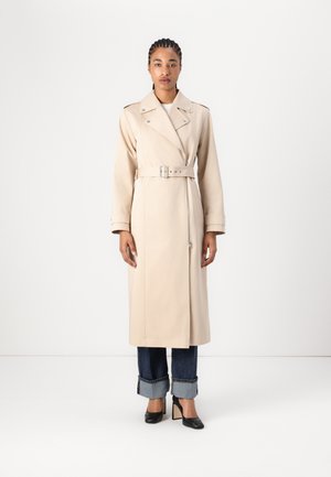 Beige Trenchcoat mit einem taillierten Schnitt, breiten Revers und langen Ärmeln. Der Stoff wirkt glatt und ist passgenau, kombiniert mit gekrempelten blauen Jeans und hohen Schuhen.