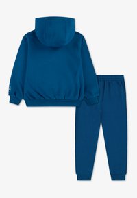 Modra bombažna kapuca s kapuco in rebrastimi manšeti, v paru s modrimi joggerji z elastičnim pasom. Mehak tekstura, preprost dizajn.