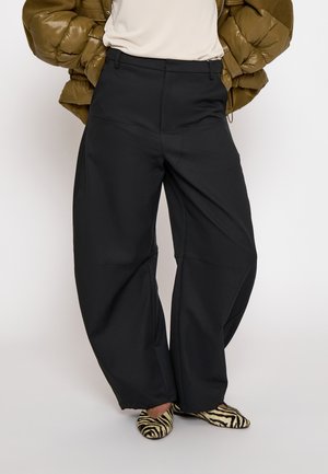 Person trägt weite schwarze Hosen, beige Oberteil, olivgrüne Daunenjacke und flache Schuhe mit Zebramuster und steht vor einem schlichten Hintergrund.