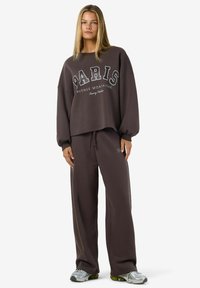 Giovane donna in piedi, con una felpa larga di colore marrone scuro e pantaloni a gamba larga abbinati, indossa sneakers argentate su sfondo bianco.