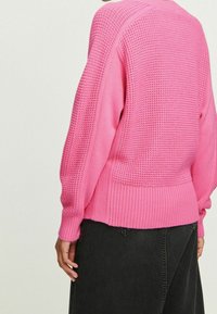 Person, der einen strukturierten pinken Strickpullover und einen schwarzen Jeansrock trägt, von hinten gezeigt, mit einer entspannten Hand an der Seite.