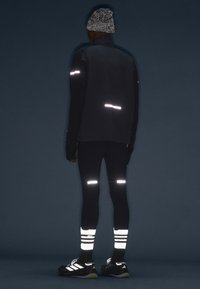 Black reflective running jacket and tights, featuring horizontal reflective stripes. Black sneakers with white accents and striped socks.