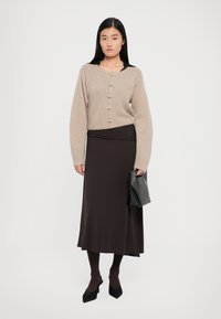 Beige knitted cardigan with large buttons, black top underneath, long brown skirt, black handbag, and black heels. Smooth textures throughout.