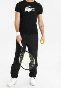 Maglietta nera a maniche corte con logo coccodrillo bianco, abbinata a pantaloni sportivi neri e sneakers bianche, mentre tiene una racchetta da tennis e una palla.