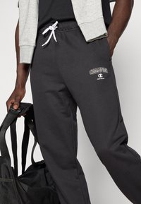 Μαύρο παντελόνι φόρμας με κορδόνι στη μέση, διακοσμημένο με λευκό κέντημα λογότυπου "Champion" στον μηρό. Κατασκευασμένο από απαλό ύφασμα, με άνετη εφαρμογή.