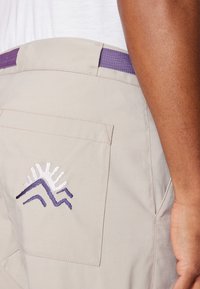 Gaiši beigi šorti ar purpura krāsas elastīgu jostu. Iekļauj aizmugurējo kabatu ar baltu un purpura krāsas kalnu masīva izšuvumu. Gluda tekstūra.