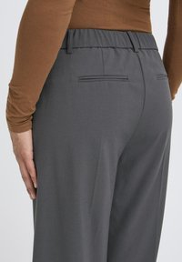 Šedé nohavice s hladkou textúrou, elastickým pásom a dvoma zadnými vreckami. Dizajn je strihaný a obsahuje minimálne kovové prvky.