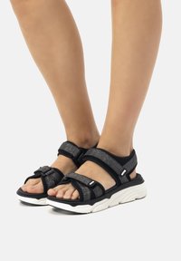 Sandalias negras con dos correas ajustables de velcro y parte superior texturizada. Presenta una suela blanca acolchada para soporte y comodidad.