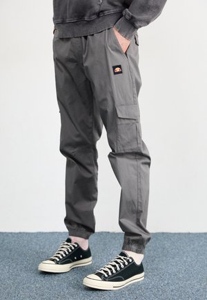 Person trägt graue Cargohosen mit elastischen Bündchen und schwarze Sneakers mit niedrigem Schaft, steht auf einem blauen Teppichboden.