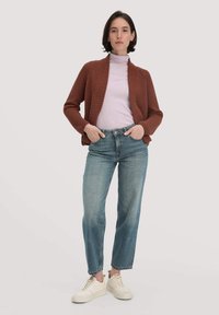 Ein rostfarbener Cardigan über einem hell-lila Rollkragenpullover, kombiniert mit hellblauen Jeans und weißen Sneakers, vor einem neutralen Hintergrund stehend.