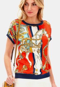 Czerwona bluzka z krótkim rękawem, zdobiona kolorowym nadrukiem koni, złotymi akcentami i ciemnogranatowym wykończeniem wzdłuż dekoltu i hemu. Gładka faktura materiału.