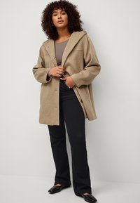 Beige, wollen blend jas met een capuchon, knoopsluiting en een ontspannen pasvorm, gecombineerd met zwarte flared broek en platte schoenen. Textuur van de stof zichtbaar.