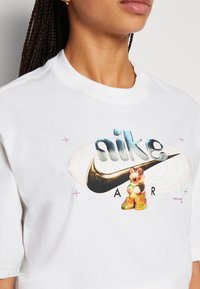 Person som bär en vit T-shirt med en stiliserad "nike air"-logotyp, svart swoosh och en färgglad gummy bear-grafik på framsidan.