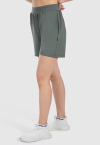 Mörkgröna sportshorts med elastisk midja, sidofickor och en smidig yta, i kombination med vita mesh-sneakers.