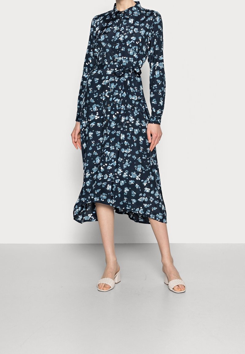 Femme portant une robe midi bleue marine à fleurs avec boutons, manches longues et ceinture à la taille, associée à des sandales blanches à talons.