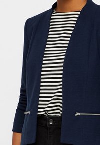 Blazer bleu marine avec une texture lisse, design à l'avant ouvert, accents zippés, porté sur un haut à rayures noir et blanc.