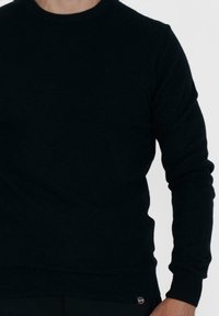 Maglione nero a collo rotondo realizzato in materiale a maglia, con maniche lunghe e una piccola etichetta con logo in basso a sinistra. Texture liscia.
