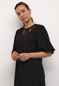 Schwarzes Kleid mit kurzen Ärmeln und einer Spitzen-V-Ausschnitt. Hat vertikale Rippungen und eine lockere Passform. Das Model zeigt die Hände zusammengehalten.