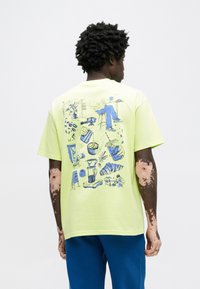 Bavlnené tričko limetkovo zelenej farby s modrou grafickou ilustráciou na chrbte, zobrazujúcou rôzne predmety a scény súvisiace s kávou.