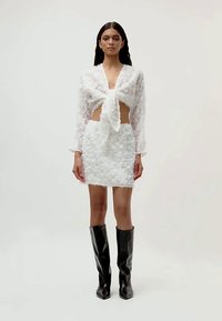 Femme portant un ensemble à deux pièces texturé blanc avec un haut noué à l'avant et une minijupe, associé à des bottes noires jusqu'aux genoux, se tenant devant un fond uni.