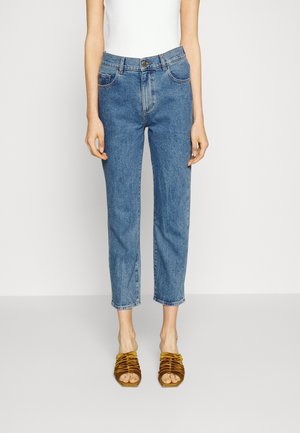 Personne portant un jean droit bleu taille haute et un haut blanc sans manches avec des talons ouverts marron froncés sur un fond uni.