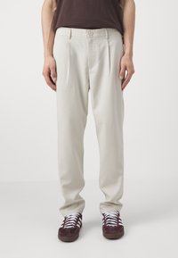 Pantalons beige clair avec plis, en tissu lisse. Dotés d'une fermeture à bouton et de poches latérales. Associés à des baskets rouge foncé.