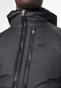 Svart puffjacka med hög krage, dubbla dragkedjor och en diskret Nike-logotyp på bröstet. Har en slät, matt textur.