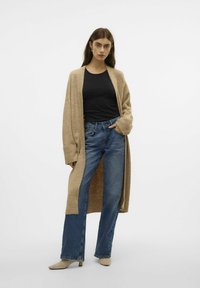 Beige lange Strickjacke über einem schwarzen, eng sitzenden Top, kombiniert mit hellblauen Weitbeinjeans und beige farbenen, hohen Mules. Glattes Material und entspannter Schnitt.