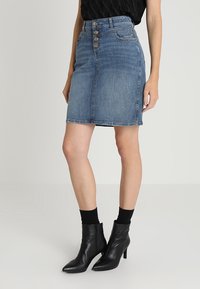 Falda lápiz de denim con cintura alta, cierre de cinco botones y un lavado azul medio. Se lleva con botas negras hasta el tobillo y calcetines.