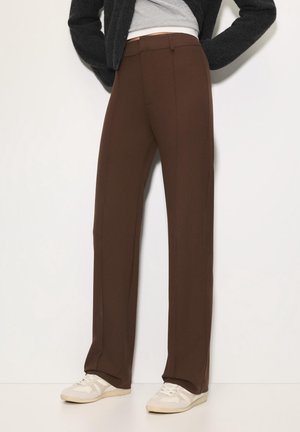 Person trägt braune, gerade geschnittene Hose mit hohem Bund, weiße Turnschuhe, graues Oberteil und schwarze Jacke, steht vor einer schlichten weißen Wand.