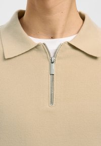 Polo di maglia beige con mezza cerniera argentata parzialmente aperta, indossata sopra una maglietta bianca, mostrando la zona del collo e del colletto.