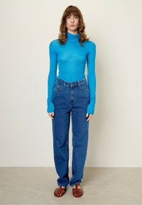 Turquoise rekbare lange mouw turtleneck, gecombineerd met hoge taille blauwe jeans en bruine gevlochten sandalen. Eenvoudige snit en pasvorm.