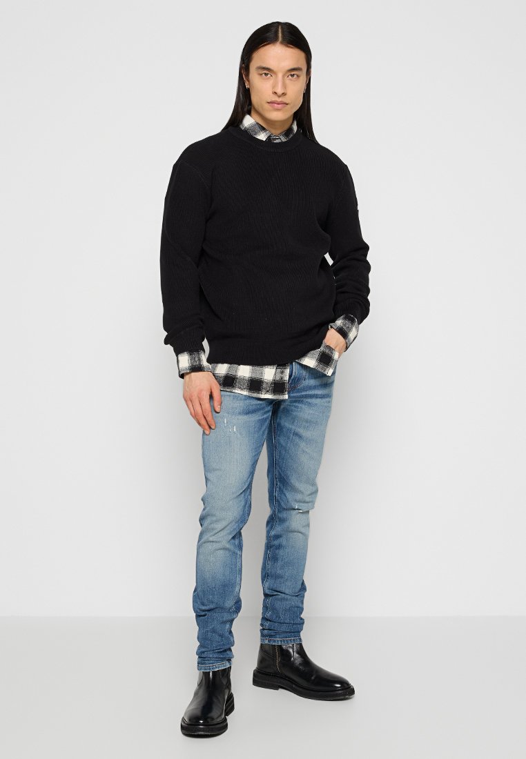 Maglione nero a costine indossato sopra una camicia a quadri bianco e nera, abbinato a jeans chiari strappati e stivali alla caviglia in pelle nera.